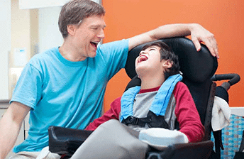 A father sits next to his son with cp who is sitting in his wheelchair. Both are smiling at each other