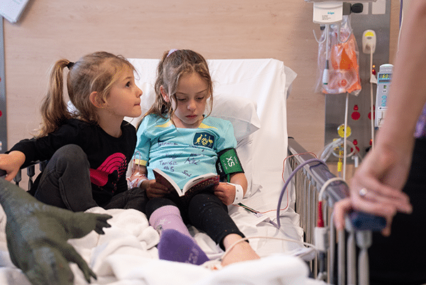 Zara sitting in a hospital bed reading a book with her younger sister next to her, while she receives her umbilical cord blood infusion.