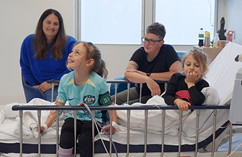 Zara sitting on a hospital bed smiling while she receives her umbilical cord blood infusion, with her younger sister and her parents around her.