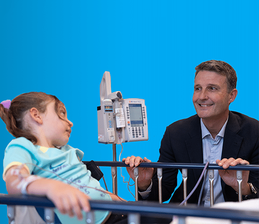A young girl sitting on a hospital bed during umbilical cord blood treatment, with a male doctor next to her, talking and smiling with her.