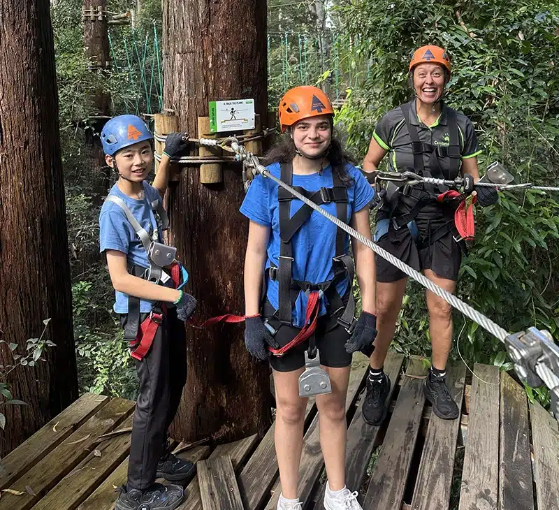 Two children and a therapist wearing harnesses and helmets, standing on a platform in the bush at treetops on the central coast