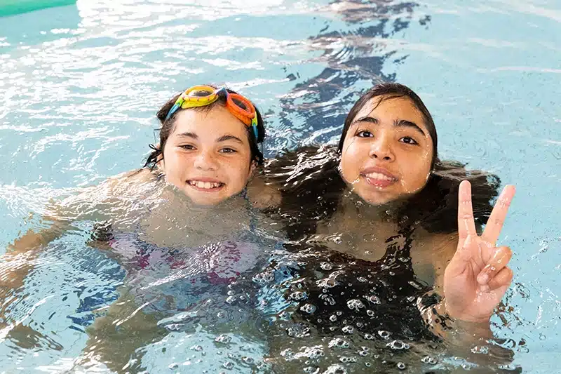 Two young girls in a swimming pool, standing close together with their heads out of water, smiling to camera.