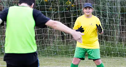 A young girl standing in a soccer goal ready to defend while another player takes a shot at goal towards her