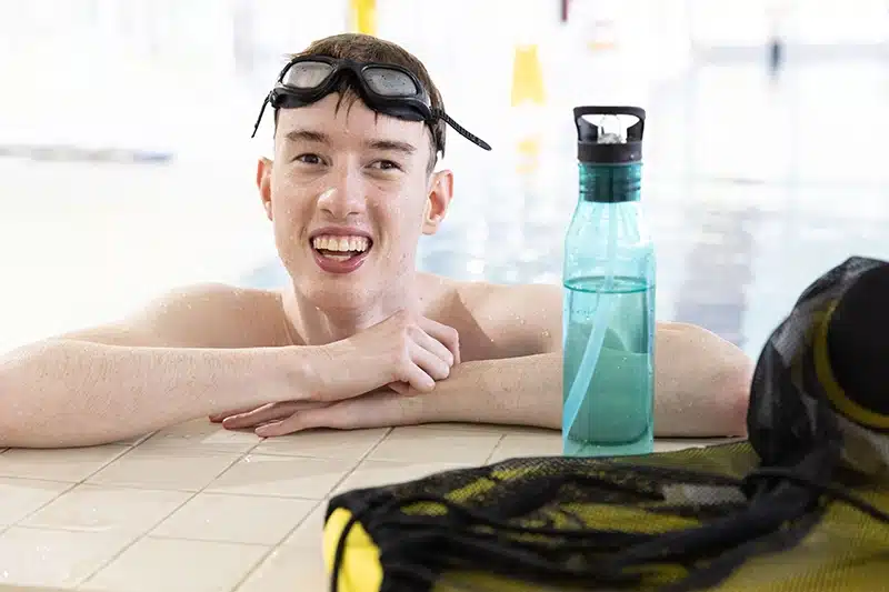 A young adult male standing in a swimming pool, resting his arms on the ledge, smiling to camera with goggles on his head and a drink bottle next to him