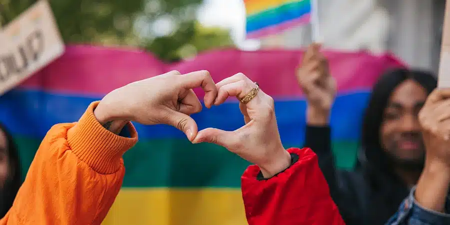 Two different hands making a heart shape against a rainbow flag celebrating love during a Pride Parade.