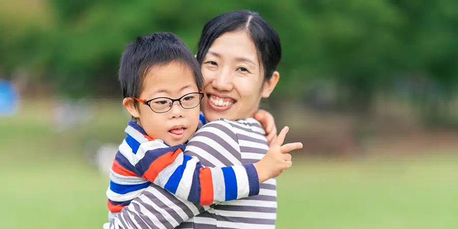 An Asian mother smiling to camera while hugging her young son with down syndrome