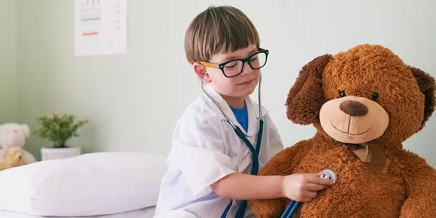 A little boy dressed up as a doctor holds a stethoscope to a large teddy bears chest