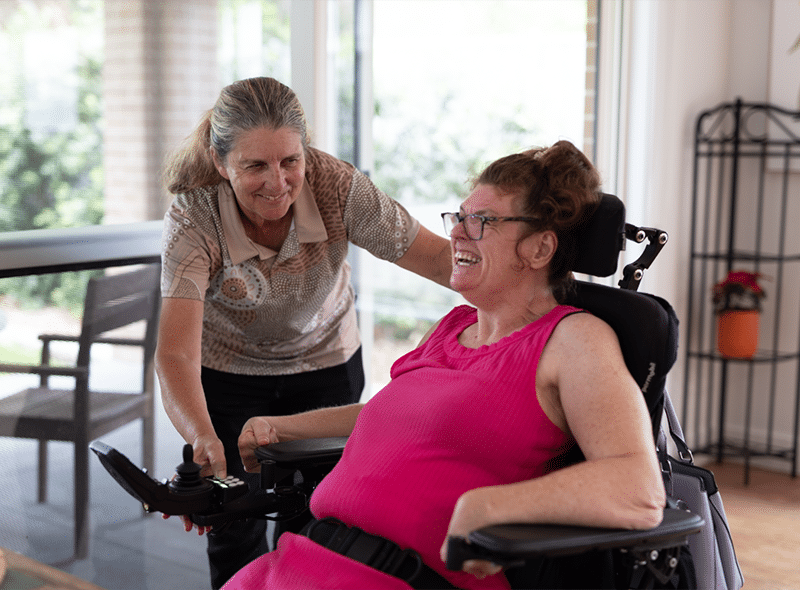A caregiver smiles and speaks with a woman in a wheelchair, gently holding the chair while the woman laughs and uses a control joystick in a bright living room.