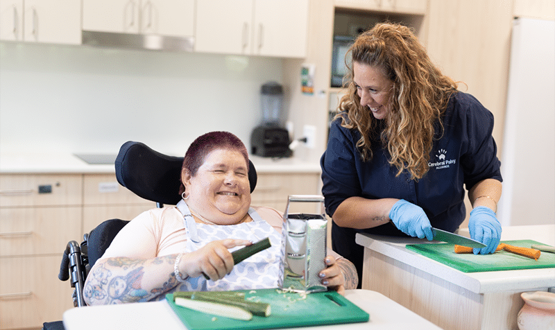 A woman in a wheelchair smiles while grating a cucumber at a kitchen counter, assisted by a caregiver who is cutting vegetables beside her.