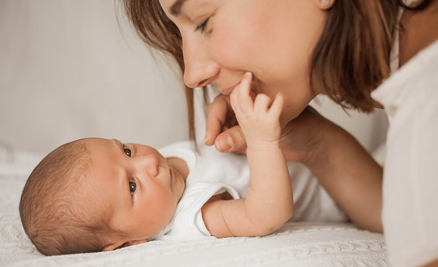 Newborn baby lying on a white blanket and gently holding a caregiver's finger during a close-up bonding moment.