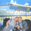 Maria and Lindsay smiling together in their wheelchairs in front of an image of a large cruise ship on the water.
