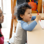 A young boy standing at a table holding a pencil with a therapist sitting behind him watching on