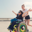 A girl in a wheelchair smiling with her arms outstretched with a friend behind her pushing the wheelchair and smiling
