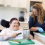 A woman in a wheelchair smiles while grating a cucumber at a kitchen counter, assisted by a caregiver who is cutting vegetables beside her.
