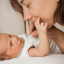 Newborn baby lying on a white blanket and gently holding a caregiver's finger during a close-up bonding moment.