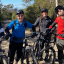 A group of 4 people in bike riding attire, standing together with their bikes in the bush