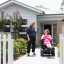 A support worker stands next to a woman in a wheelchair, smiling to each other in the front garden of a brick house