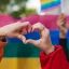 Two different hands making a heart shape against a rainbow flag celebrating love during a Pride Parade.