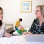 A mother and therapist having a discussion at a table while her young son plays with toys in the background
